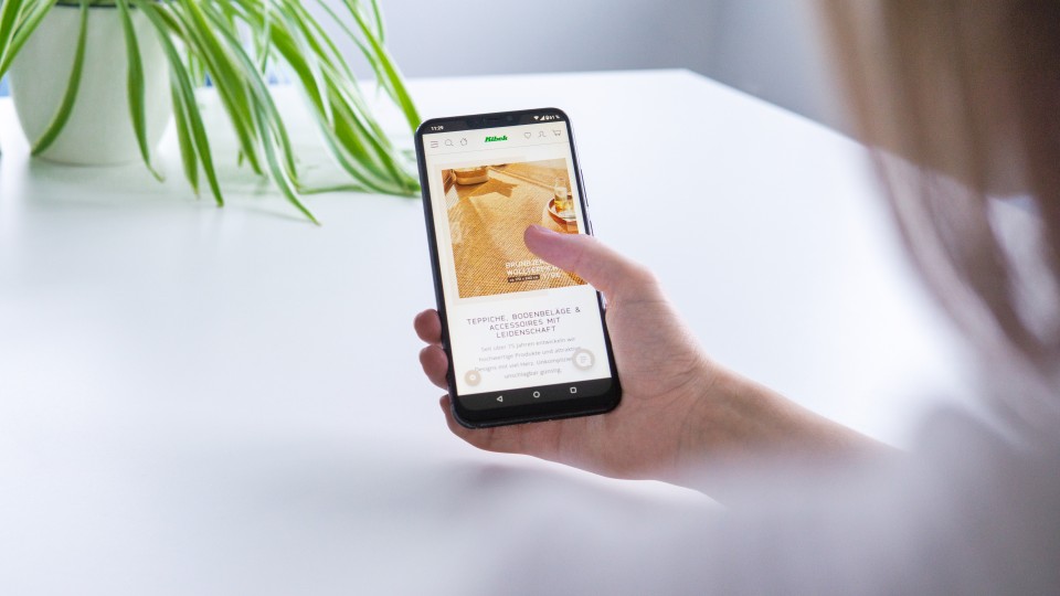 Jemand zeigt auf dem Smartphone den Webshop des Teppichhändlers Kibek