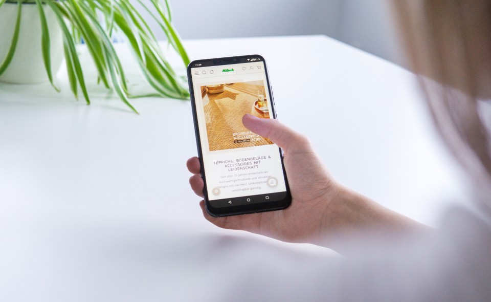 Jemand zeigt auf dem Smartphone den Webshop des Teppichhändlers Kibek