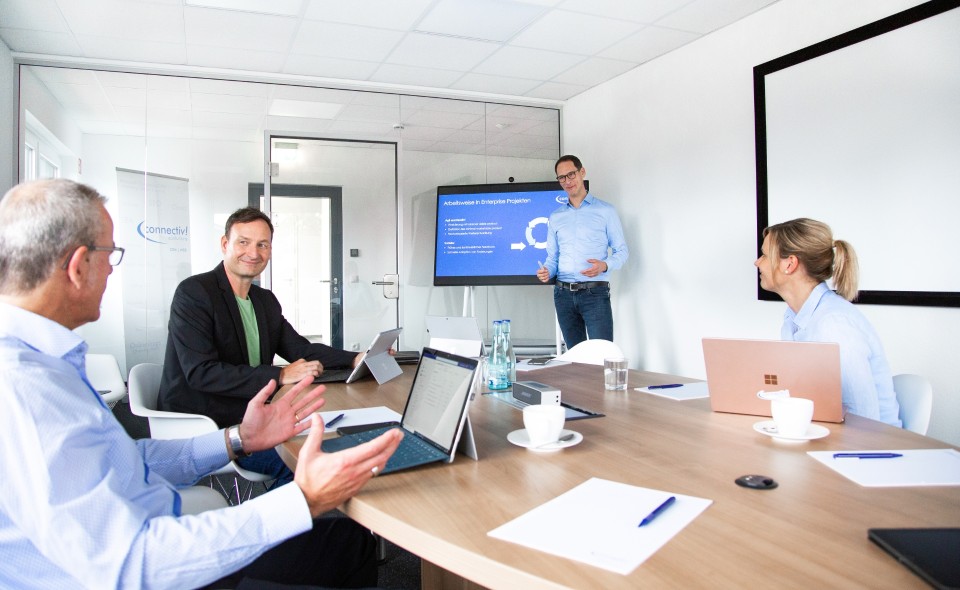 Drei Personen sitzen am Tisch und eine weitere präsentiert etwas am Surface Hub